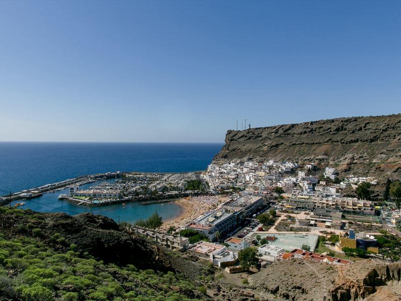 Paisaje costero con cielo azul claro, mar y un pequeño puerto frente a una montaña rocosa.