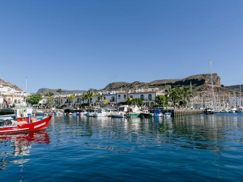 Puerto deportivo con barcos frente a un pequeño pueblo y colinas al fondo bajo cielo despejado.