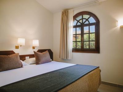 Chambre d'hôtel simple avec lit double, coussins, murs clairs et fenêtre ronde avec rideau.