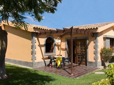 Kleines Ferienhaus mit Terrasse, Stühlen und Tisch in sonniger, grüner Umgebung.