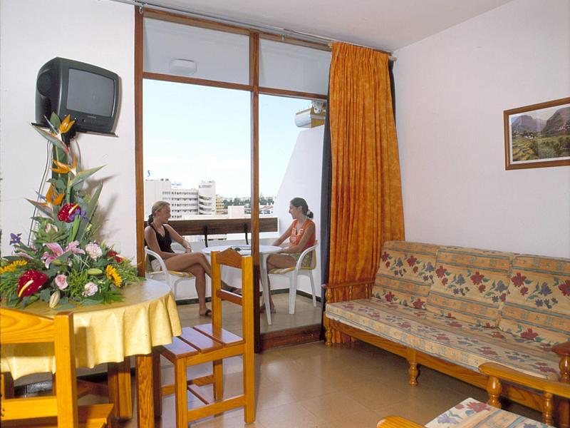 Habitación de hotel con balcón, dos personas sentadas afuera, flores coloridas y mobiliario sencillo.