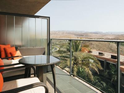 Balkon mit Glasgeländer, Sitzbereich mit orangen Kissen, Blick auf Palmen und Hügel im Hintergrund.