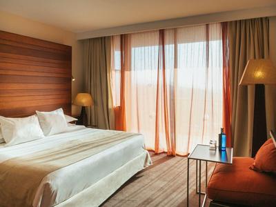 Hell eingerichtetes Schlafzimmer mit großem Bett, braunen Vorhängen und gemütlichem Sessel.