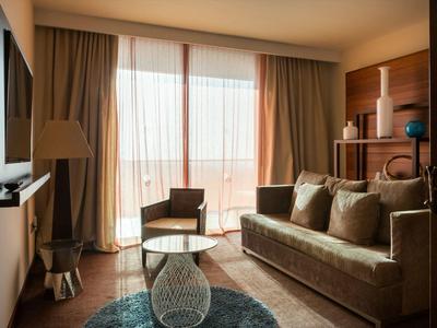 Gemütliches Wohnzimmer mit braunem Sofa, Glas-Couchtisch und großen Fenstern mit Vorhängen.