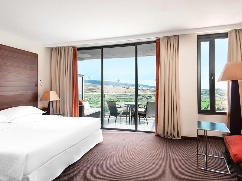 Geräumiges Hotelzimmer mit großem Bett, Balkon mit Meerblick, roten Sesseln und Holzvertäfelung.