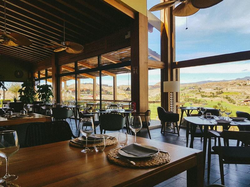 Gemütliches Restaurant mit Holztischen, schwarzen Stühlen und weitem Fensterblick auf grüne Landschaft.