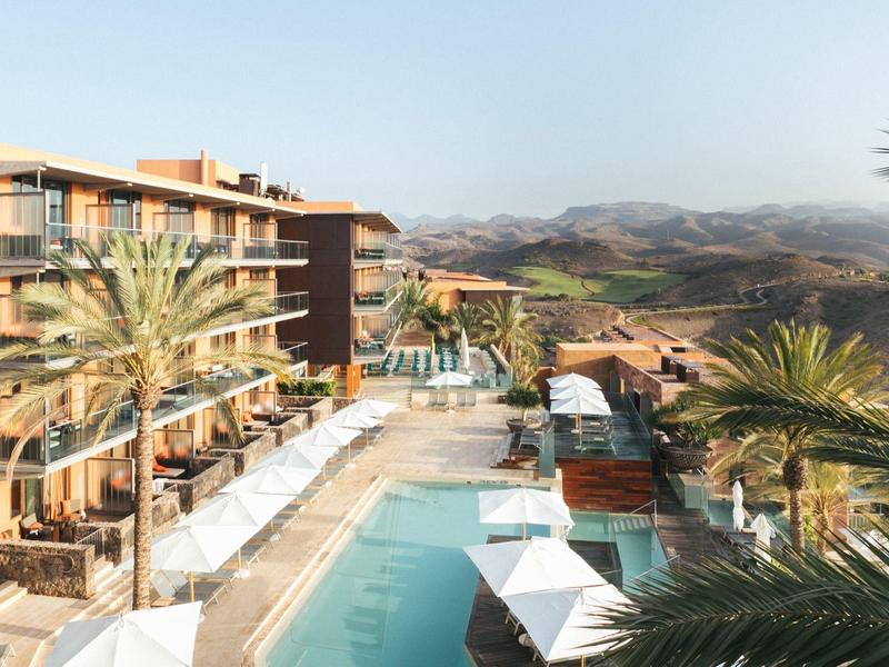 Hotel mit Pool, Palmen und Terrasse vor bergiger, grüner Landschaft unter klarem Himmel.