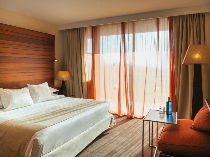 Hell eingerichtetes Schlafzimmer mit großem Bett, braunen Vorhängen und gemütlichem Sessel.