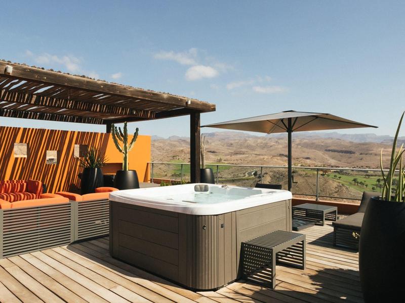 Moderner Outdoor-Whirlpool auf Holzterrasse mit Sitzbank, Sonnenschirm und weitem Landschaftsblick.