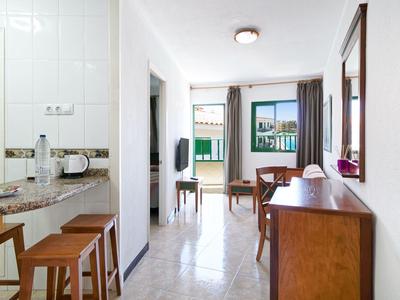 Camera d'hotel luminosa e semplice con angolo cottura, tavolo da pranzo, sedie e balcone con vista sul verde.