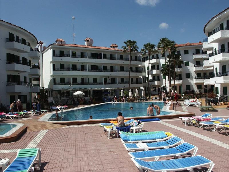 Großer Pool mit blauen Liegen vor mehrstöckigem, weißem Hotel unter blauem Himmel.