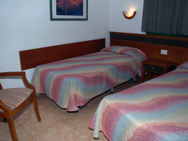 Einfaches Zimmer mit zwei Einzelbetten, bunten Decken, Holzmöbeln und einem Bild an der weißen Wand.