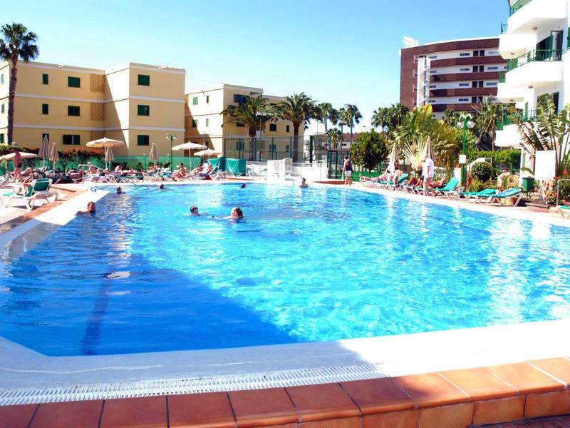 Großer blauer Pool mit Sonnenschirmen, Liegestühlen und mehrstöckigen Gebäuden im Hintergrund.