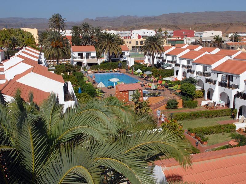 Hotelcomplex met rode daken, zwembad en palmbomen in een zonnig landschap.