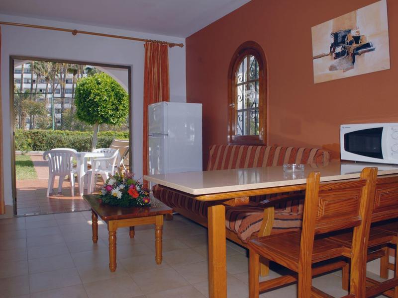 Eetgedeelte met houten tafel, stoelen, bank, koelkast en toegang tot terras met tuinmeubilair.