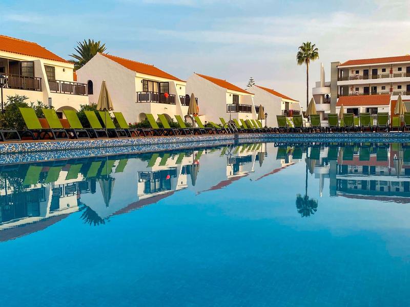 Groot hotelzwembad met ligstoelen en witte gebouwen met rode daken op de achtergrond.