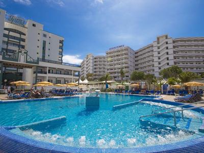 Großer Hotelpool mit Wasserspielen, Sonnenliegen und mehrstöckigen Gebäuden im Hintergrund.