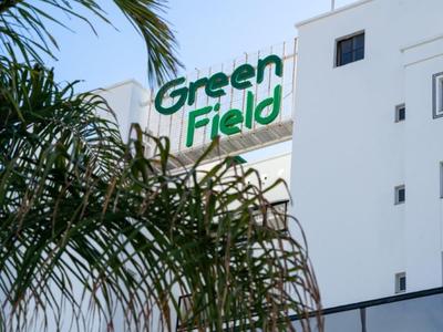 Moderner Hotelbau mit sichtbarem grünem "Green Field" Schriftzug und Palmen im Vordergrund.