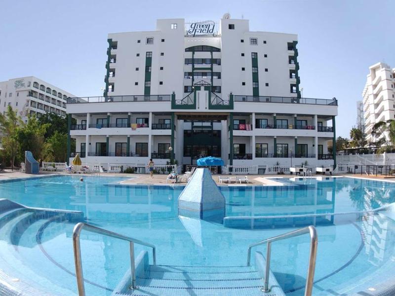 Modern hotel with large outdoor pool in sunny location, surrounded by multiple buildings.