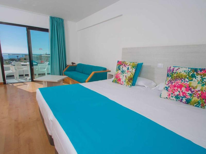 Hotel room with large bed, colorful pillows, and balcony with sea view.
