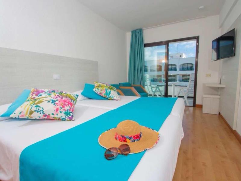 Bright hotel room with two beds, colorful pillows, and balcony with glass front.