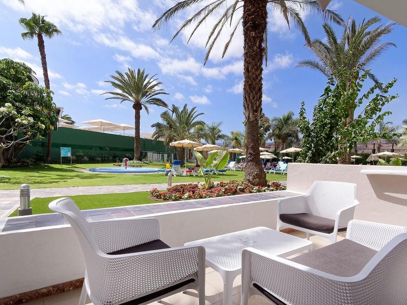 Terrasse mit weißen Stühlen, Palmen und blauem Himmel, grüner Garten im Hintergrund.