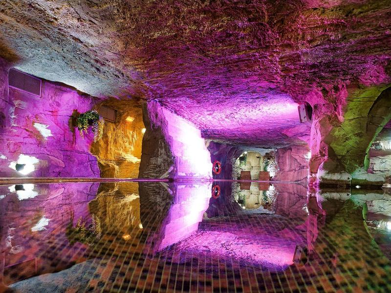 Unterirdischer Raum mit Wasserbecken, beleuchtet in Violett- und Gelbtönen, steinerne Wände und Decke.