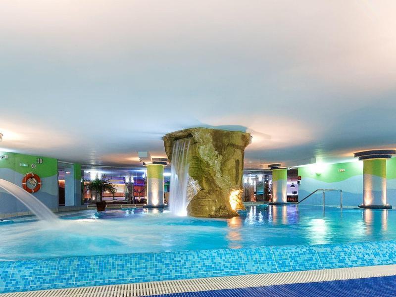 Großes Hallenbad mit Wasserfällen, Sprudelperlen und runden Säulen in hellem Ambiente.