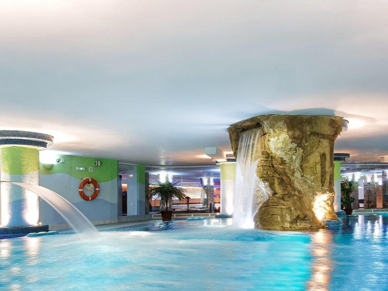 Hallenbad mit blauem Wasser, Wasserrutsche, Felsenwand und weißen Säulen unter heller Decke.