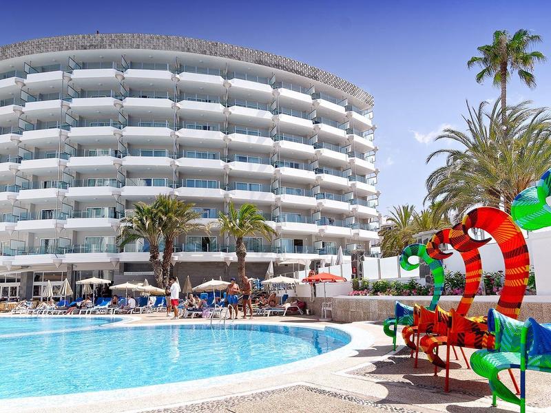 Rundes Hotel mit weißen Balkonen, blauem Pool und bunten Sonnenschirmen in tropischer Umgebung.