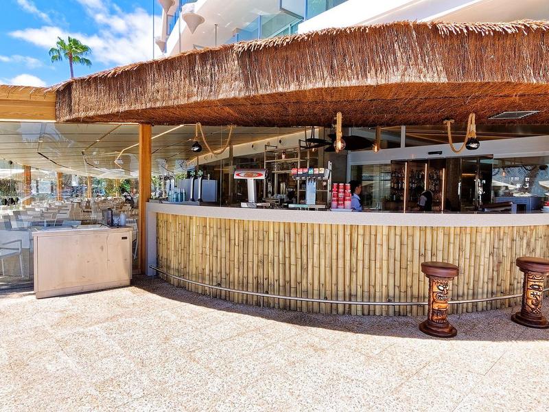 Offene Bar mit Bambusverkleidung, Strohdach und Holzstühlen auf heller Terrasse vor Hotel.