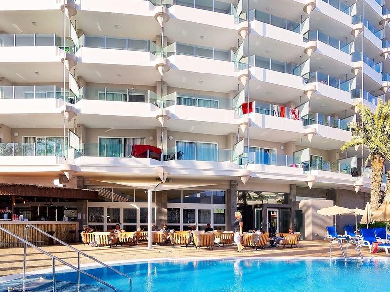 Moderner Hotelkomplex mit mehreren Balkonreihen, Außenpool, Liegestühlen und Palmen bei klarem Himmel.