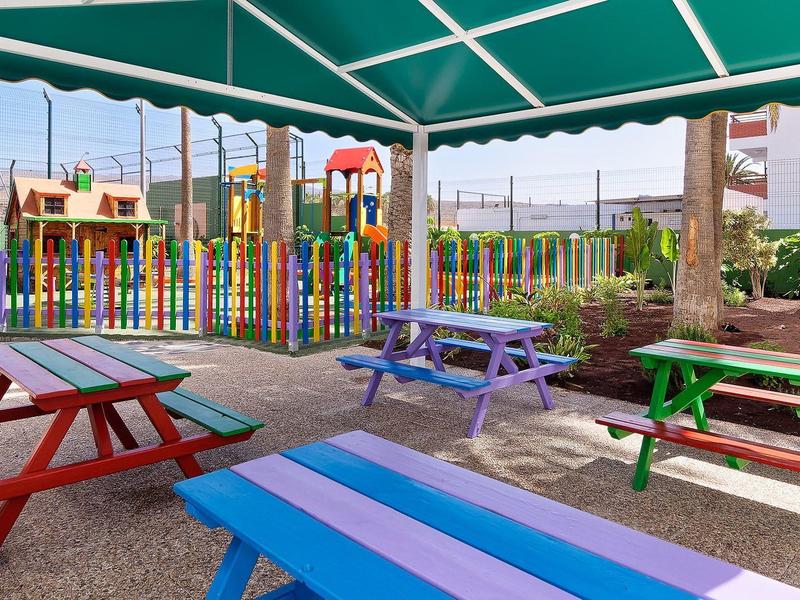 Bunte Bänke und Tische unter grünem Sonnendach mit buntem Spielplatz im Hintergrund.