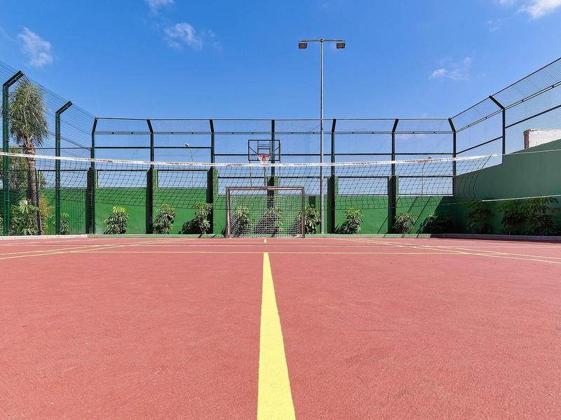 Blick auf einen roten Tennisplatz mit gelber Mittellinie, umgeben von hohem Zaun und blauem Himmel.