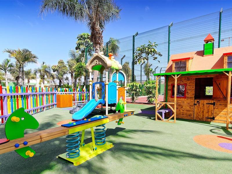 Bunter Spielplatz mit Rutsche, Wippe und Holzhaus, umgeben von Palmen und bunter Umzäunung.