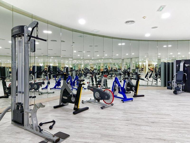 Großer moderner Fitnessraum mit Spiegelwand, vielfältigen Trainingsgeräten und hellgraßem Boden.