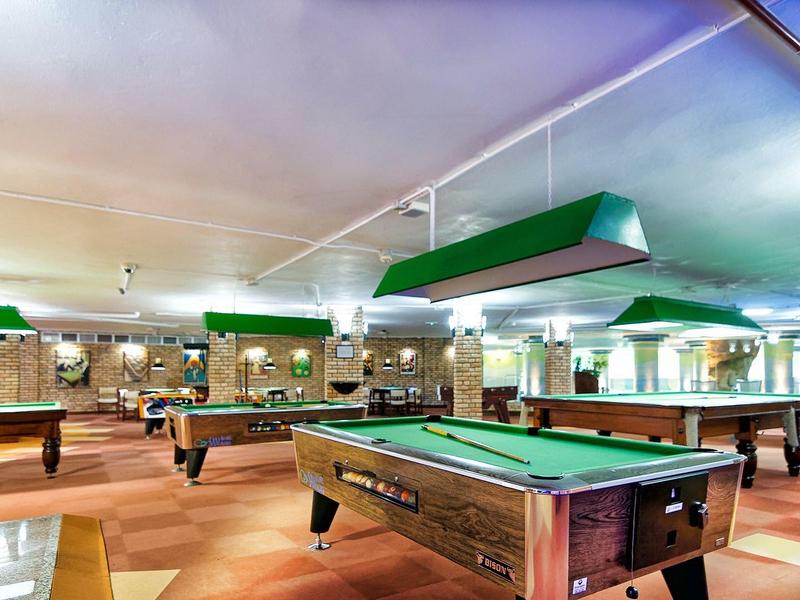 Großer heller Raum mit mehreren Billardtischen, Holzboden und grünen Lampen über den Tischen.