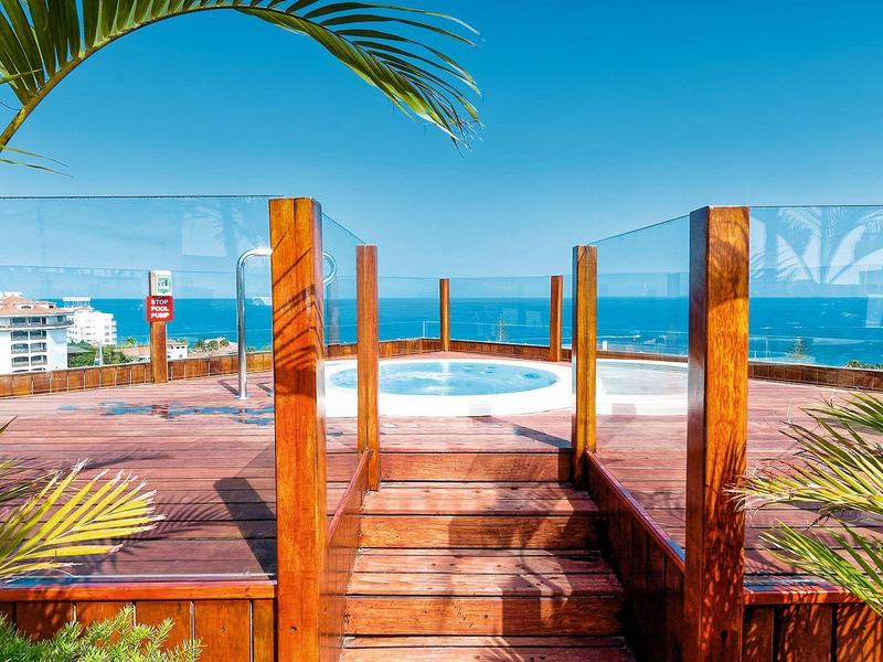 Holzdeck mit Treppe, Pflanzen rechts und links, offenem Blick aufs Meer und klarem Himmel.