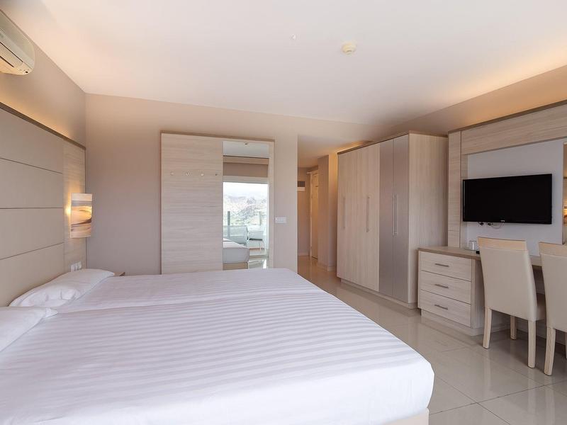 Helles Schlafzimmer mit großem Bett, Schreibtisch, Stühlen, TV und Kleiderschrank in neutralen Farben.