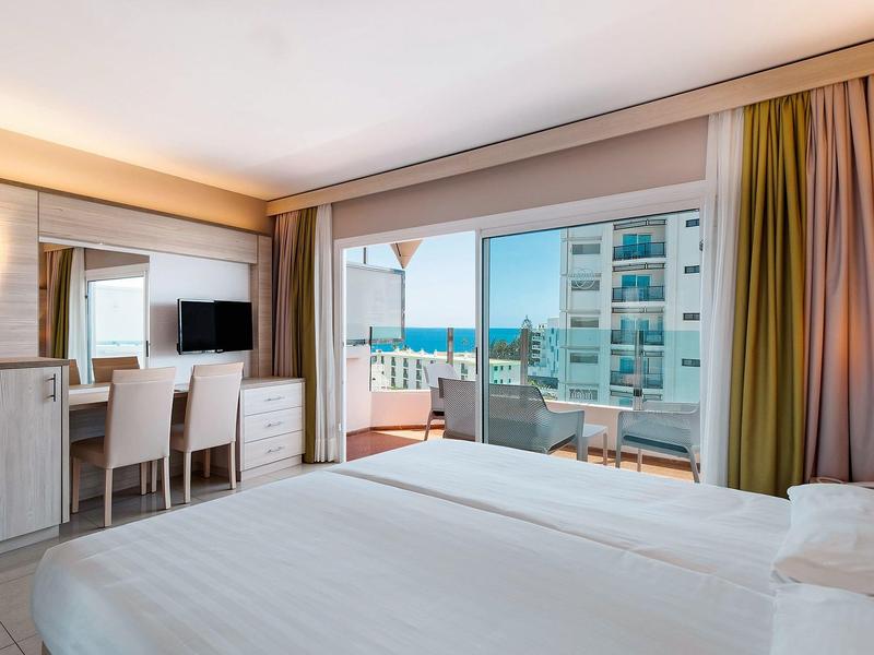 Helles Hotelzimmer mit Doppelbett, Schreibtisch, Balkon und Meerblick bei Tageslicht.