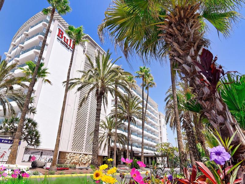 Modernes, weißes Gebäude mit runden Balkonen, umgeben von Palmen und bunten Blumen im Vordergrund.