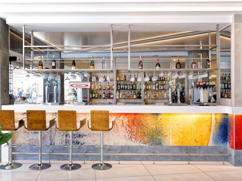 Moderner Barbereich mit fünf Hockern, bunter Thekenfront und vielen Flaschen im Regal hinter der Bar.