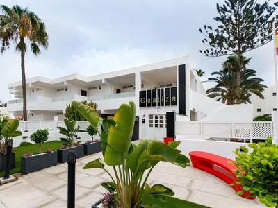 Modernes weißes Gebäude mit Balkonen, Palmen und vielfältiger grüner Bepflanzung im Vordergrund.