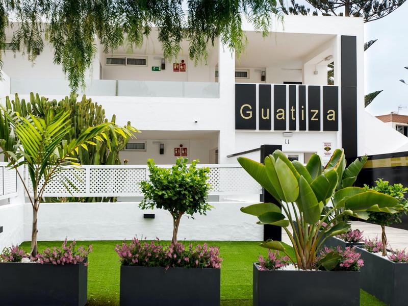 Modernes, weißes Gebäude mit großem Eingangsbereich und schwarzem Schriftzug "Guaniza". Vorgarten mit Palmen.