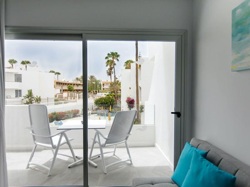 Balkon mit zwei weißen Stühlen, kleinem Tisch, Blick auf Pool und Palmen, helle moderne Wohnung.
