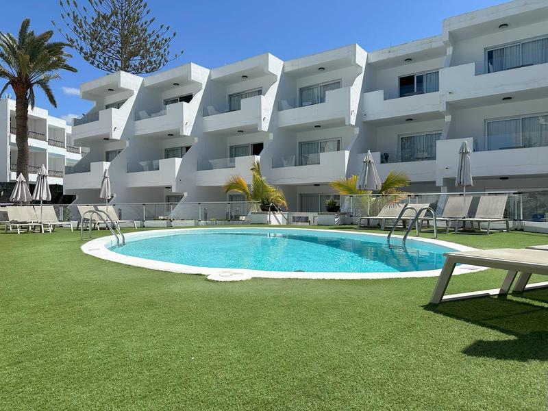 Moderner weißer Wohnkomplex mit rundem Pool und Sonnenliegen, blauer Himmel im Hintergrund.