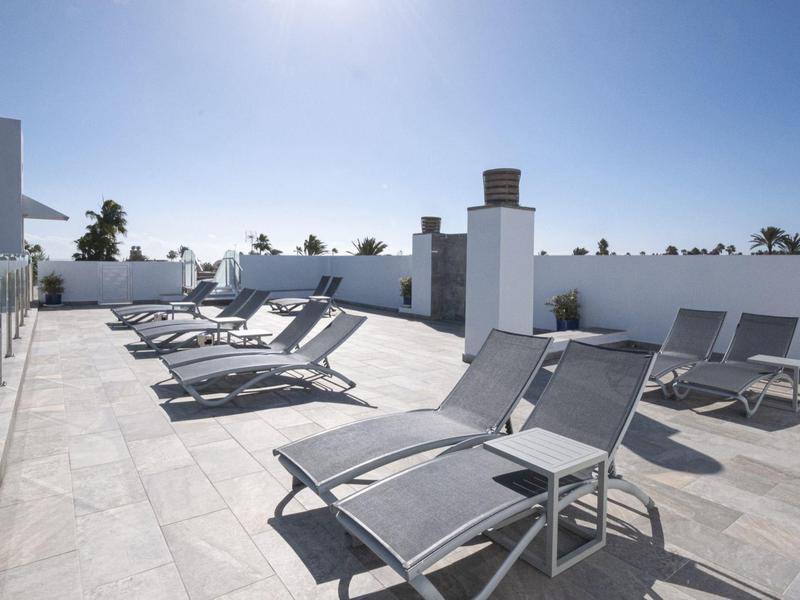 Dakterras met grijze ligstoelen en lichte vloerbedekking onder een zonnige lucht.
