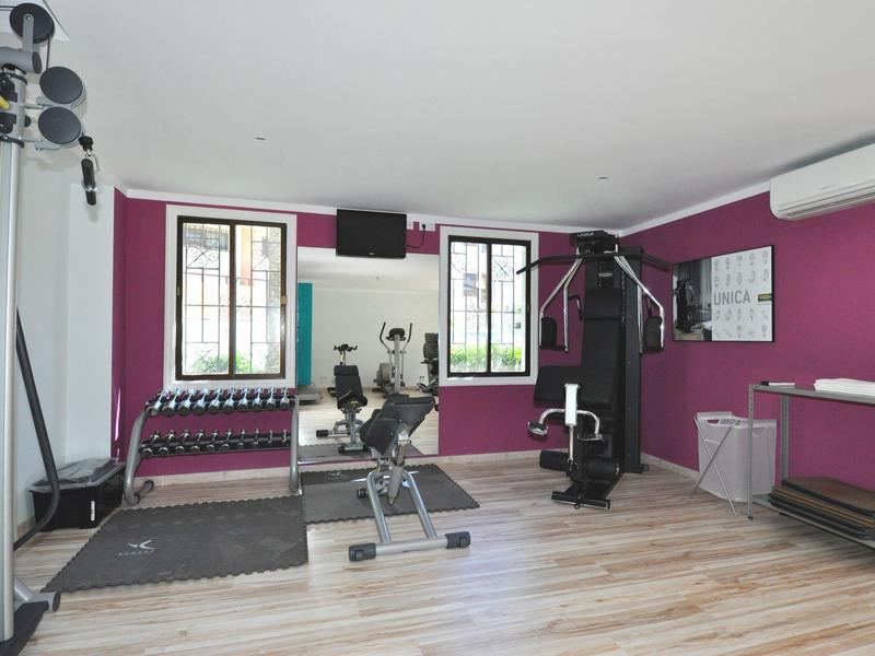 Kleine fitnessruimte met halters, krachtapparaten en twee ramen tegen een paars geschilderde muur.