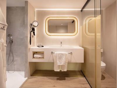 Moderní hotelová koupelna se světlým zrcadlem, umyvadlem a sprchou bez dveří.