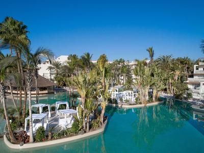 Großer Pool mit Palmen und weißen Liegestühlen unter klarem blauem Himmel in einer Hotelanlage.
